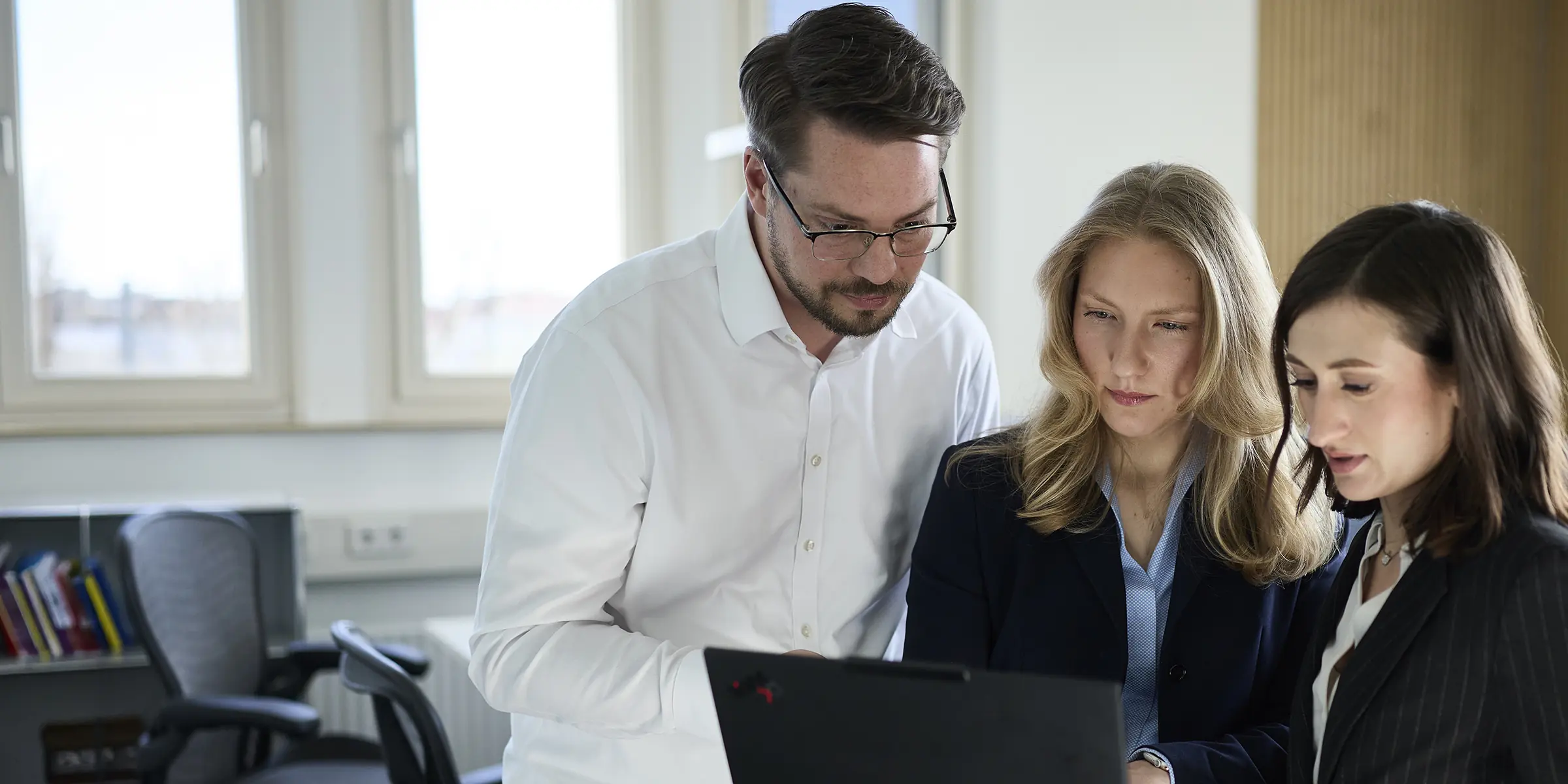 Drei Personen in Business-Kleidung schauen gemeinsam auf einen Laptop in einem hellen Büro.