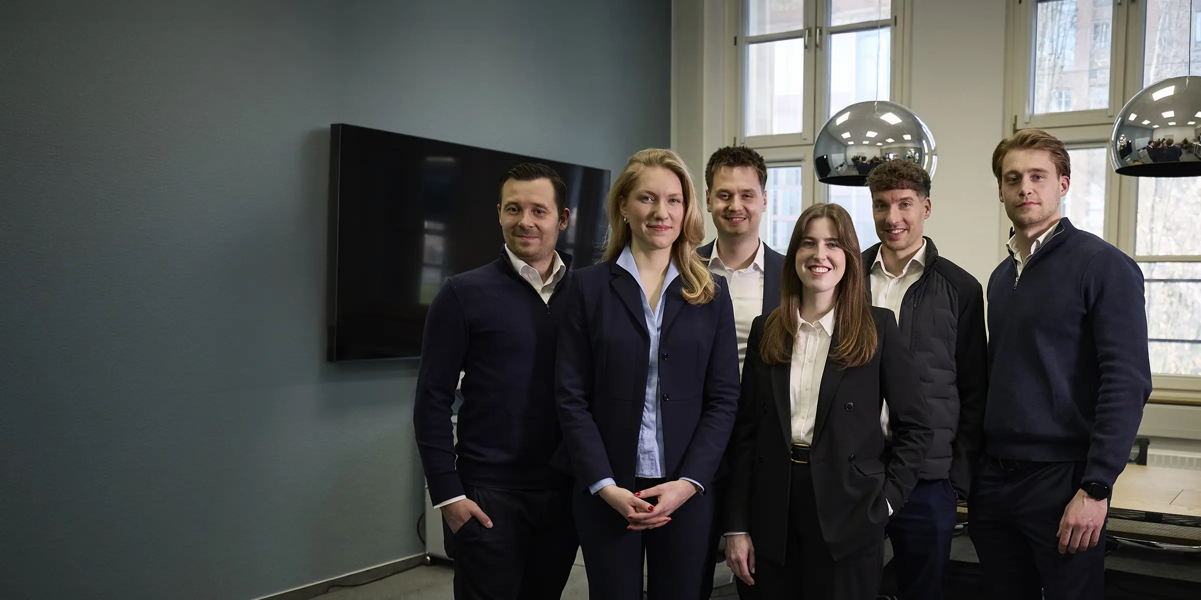 Sechsköpfiges Team in dunkler Business-Kleidung posiert lächelnd für ein Gruppenfoto in einem modernen Büro.