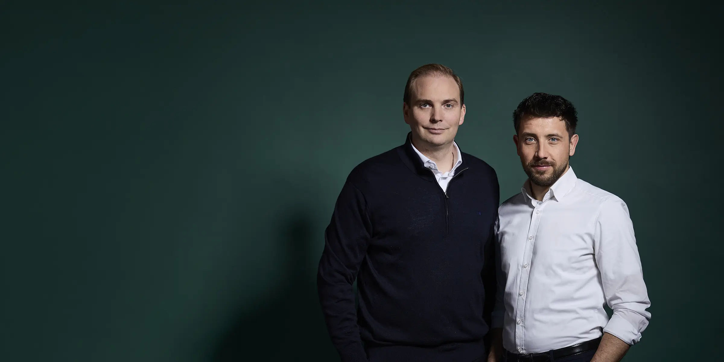 Zwei Männer in Business-Casual-Kleidung posieren lächelnd vor dunkelgrünem Hintergrund.