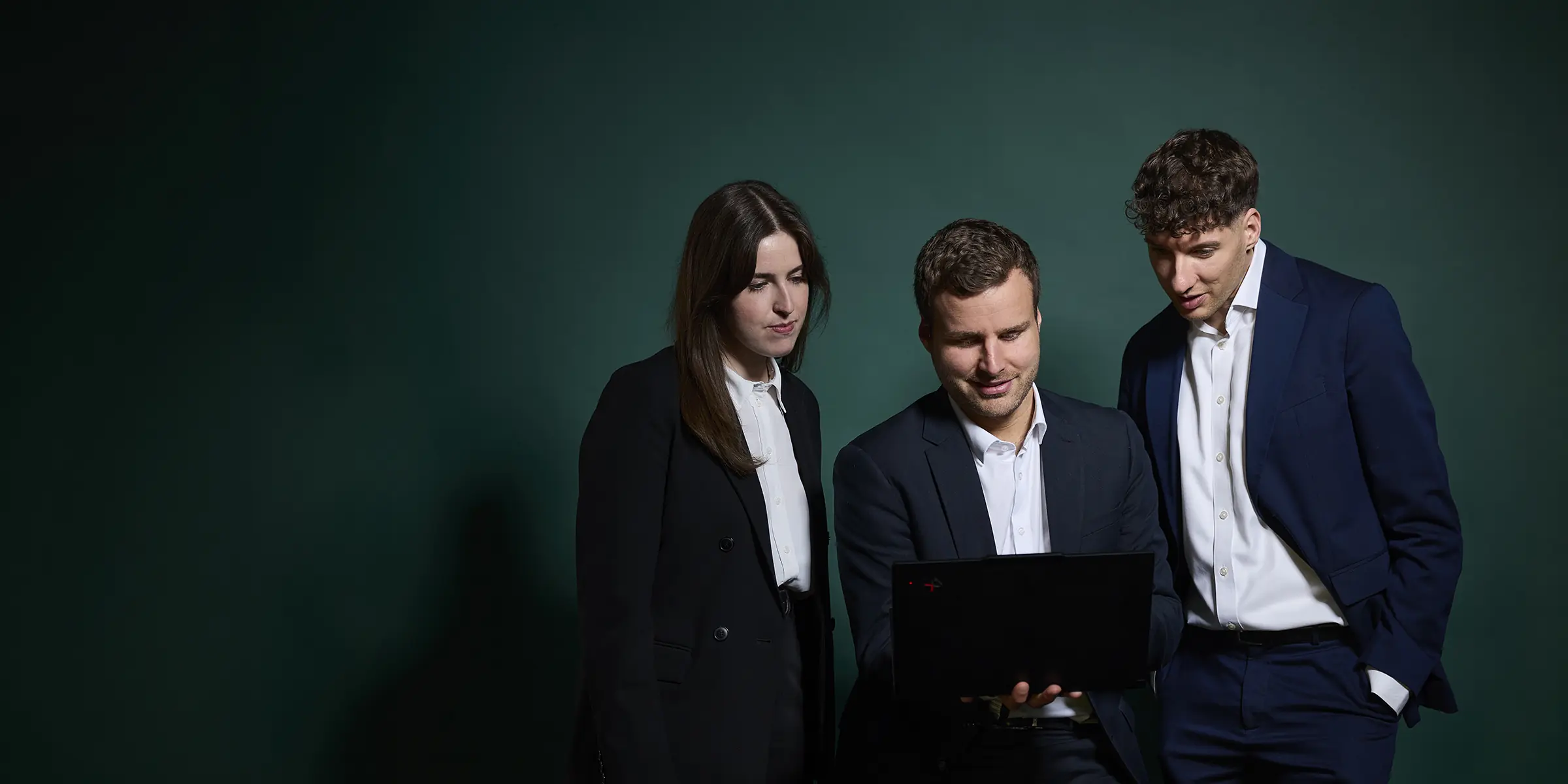 Drei Personen in dunkler Business-Kleidung schauen gemeinsam auf einen Laptop vor dunkelgrünem Hintergrund.